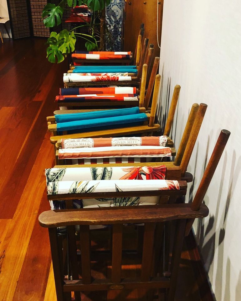 Seven brightly coloured directors chairs stand folded in a row in the Chisholm St venue. There is a monstera plant in the background and wooden floorboards.