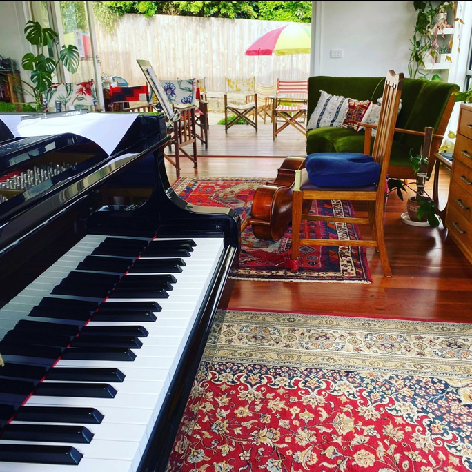 A view of the upper keys of a grand piano and beside it is a chair with a cello in front, lying on its side. There are brightly coloured director's chairs in distance, set up for the audience. Bright red and blue rugs are on the wooden floorboards under the cellist's chair.