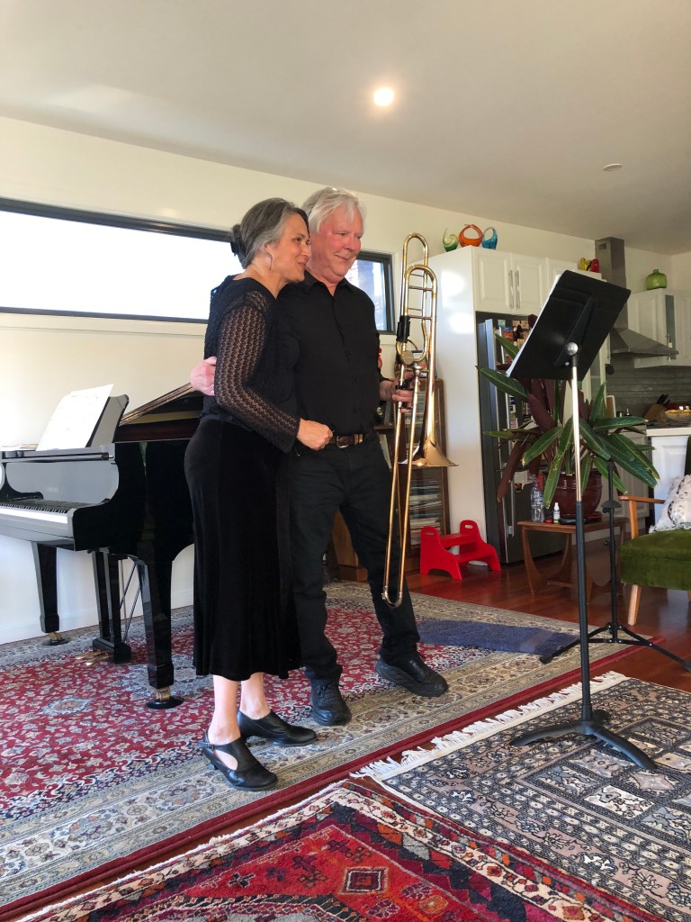 A woman dressed in black (pianist and host Bronwyn Blaiklock) stands beside a man who is holding a trombone (Hugh McKelvey) and they are both smiling. This was taken at a 2023 Chisholm St that solo trombone works.