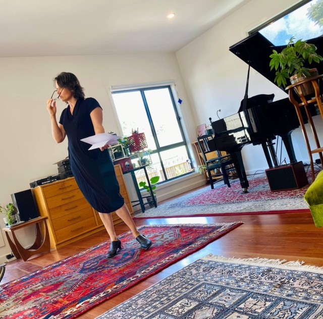 A moment taken of pianist and host, Bronwyn Blaiklock, as she removes her glasses whilst speaking to the audience. She is holding notes on paper, and the image is taken on an angle. There are red and blue rugs on the wooden floorboards and a large window in the background.