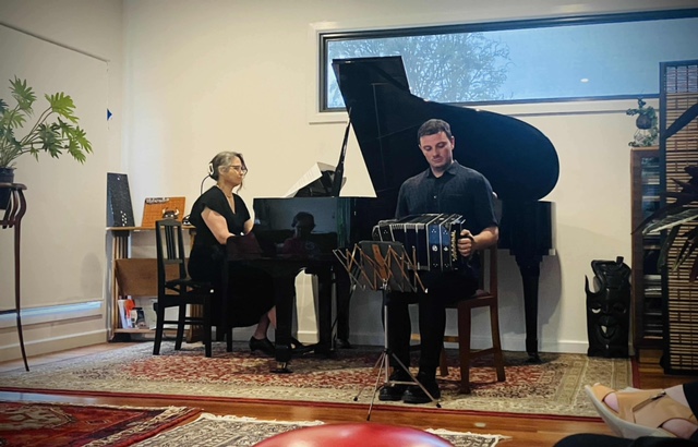 Two musicians performing in a 2024 Chisholm St concert. At the grand piano is pianist and venue host, Bronwyn Blaiklock, and seated in front of the piano, performing the bandoneon is Stephen Cuttriss.