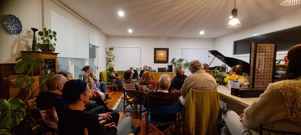 A view of the Chisholm St venue with a 'full house' inside audience: audience members are seated on colourful director's chairs, facing the black grand piano in the right corner of the room. This was taken during a 2024 concert series that featured solo piano works performed by Bronwyn Blaiklock and solo flute works performed by Sarah Goodbourn.