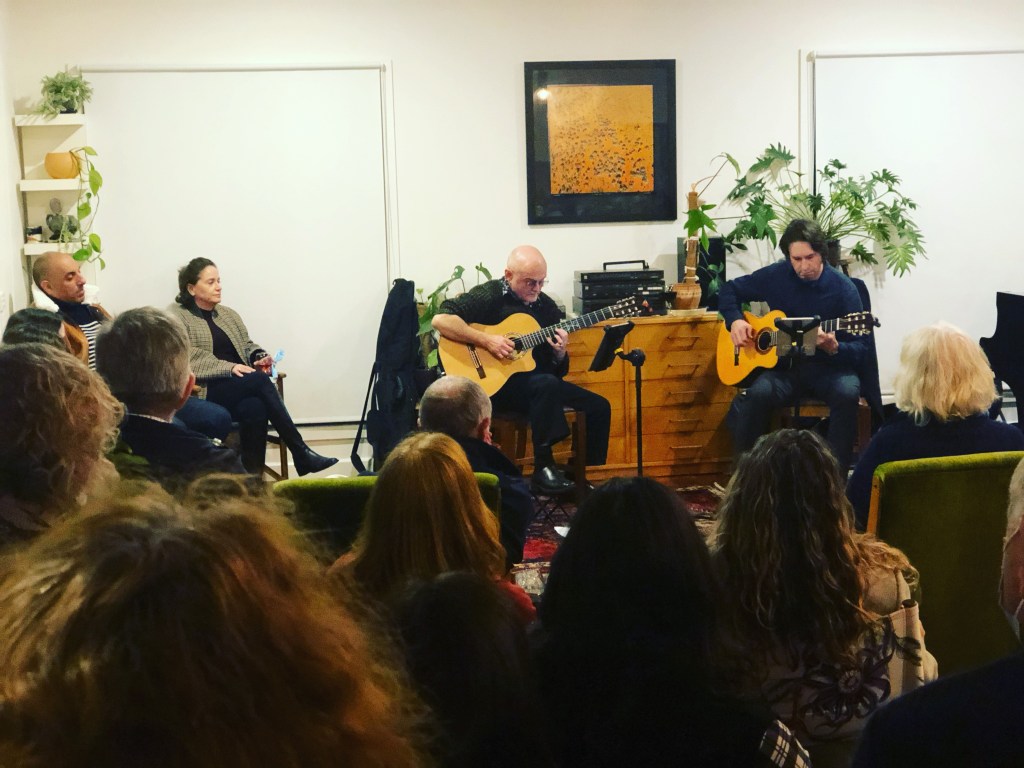 Two guitarists - Lyndon Kriss on the left, and Adam Seery on the right - perform in front of an audience. The photo was taken in a 2023 Chisholm St performance. There are potted plants around the performance space and the grand piano is the the right of the image.