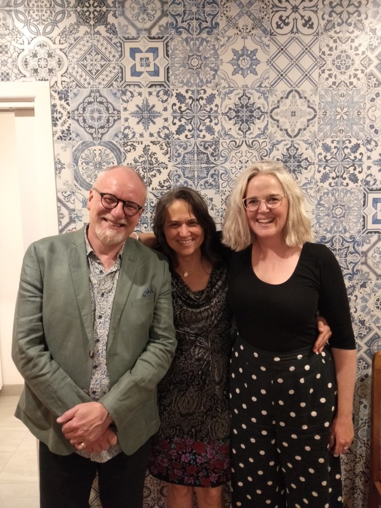 Three people stand in front of a wall decorated with blue and white tiles (left to right: Richard Chew, Bronwyn Blaiklock, and Sharon Turley). They are smiling and relaxed, and the photo was taken at Chisholm St in March, 2023.